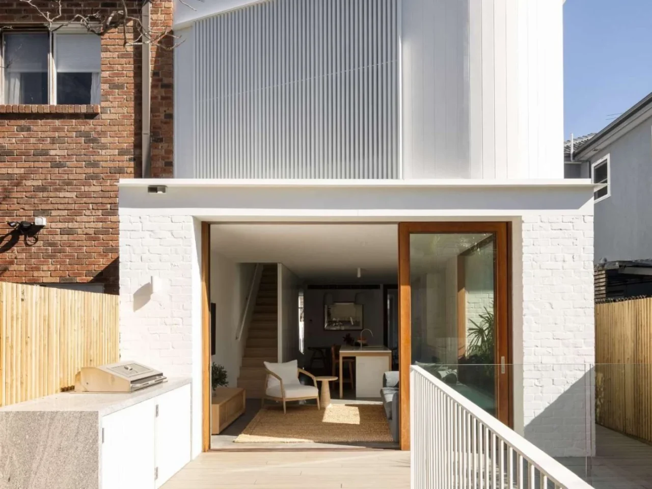 Renovated rear of an Australian brick home by Modify Homes: white upper level with vertical battens and timber-framed sliding glass doors to the deck
